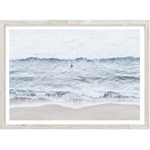 May include: A framed photograph of a lone surfer paddling in the ocean, with white capped waves crashing on the sandy beach in the foreground. The sky is a light blue, and the water is a deep blue.