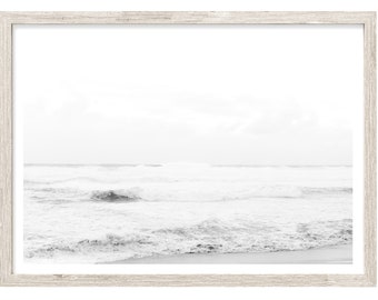 May include: A framed grayscale print of a beach scene. The image captures the ocean waves crashing onto the shore. The frame is a light, natural wood color, adding a touch of warmth to the monochromatic artwork.