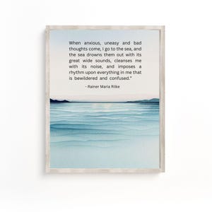 May include: A framed print with a watercolour depiction of the sea in shades of blue. The artwork includes a quote by Rainer Maria Rilke: "When anxious, uneasy and bad thoughts come, I go to the sea..." The frame is a light wood colour.