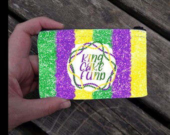 May include: A small purple, green, and yellow zippered pouch with a white circle in the center that says "King Cake Fun" in black text. The circle is surrounded by a green and purple beaded garland.
