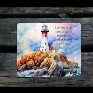 May include: A watercolor painting of a lighthouse on a rocky shore with a blue sky and white clouds. The lighthouse is red and white with a small house at the base. The text "I have come into the world as a light, so that no one who believes in me should stay in darkness John 12:46" is written in black ink on the right side of the painting.