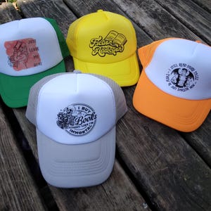 May include: Four trucker hats with different designs. The first hat is green with a white front panel featuring a red and white illustration of a person with the text "Just Leave Me Here". The second hat is yellow with a white front panel featuring the text "Fiction Addiction" and a black illustration of an open book. The third hat is orange with a white front panel featuring a black and white illustration of a person with the text "I Still Read Fairy Tales, They're Just Darker Now". The fourth hat is white with a gray mesh back and a black and white illustration of a skull with the text "I Buy Books Like I'm Immortal".