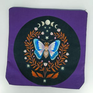 Könnte beinhalten: Lilafarbener Reißverschlussbeutel mit einem schwarzen Kreisdesign, das einen blauen und rosa Schmetterling mit einem Stern in der Mitte zeigt. Der Schmetterling ist von braunen Zweigen und weißen Blüten umgeben. Der Kreis ist von einem Halbmond und Sternen umgeben.