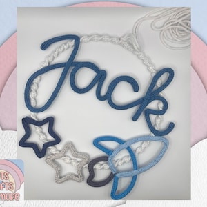 May include: A handcrafted macramé wall decoration spelling "Jack" in blue cursive lettering. It includes star and rocket ship shapes in blue, grey, and white, all suspended on a white macramé cord.