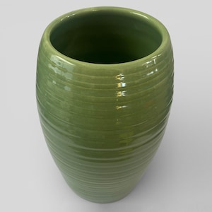 May include: A green ceramic vase with a cylindrical form and a wide opening. The vase features horizontal, ridged detailing around its exterior. The glossy finish reflects light, highlighting the vase's smooth curves. The vase is a solid, muted green colour.