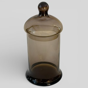 May include: A brown glass apothecary jar with a rounded lid and a spherical knob. The jar has a cylindrical body and a slightly flared base. The glass is translucent, allowing some light to pass through.