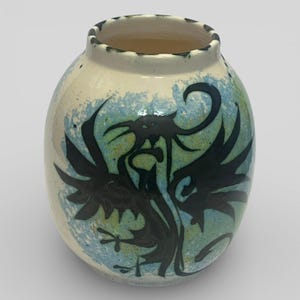 May include: A ceramic vase with a cream base and a black dragon design. The dragon is set against a blue and green background. The rim of the vase has a dark green trim.
