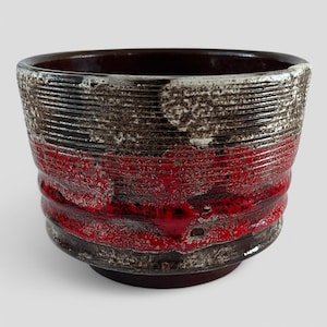 May include: A red, brown and white ceramic planter with a textured, striped design. The planter has a wide, round opening and a slightly rounded base.