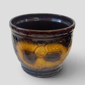 May include: A brown ceramic flower pot with a sunflower design. The pot has a dark brown rim and a lighter brown and yellow sunflower pattern on the sides. The pot is round and has a glossy finish.