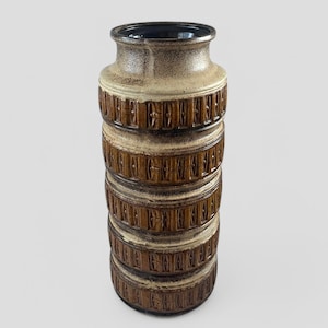 May include: A tall, cylindrical ceramic vase with a brown and cream glaze. The vase has a geometric pattern of brown lines and squares around the body.