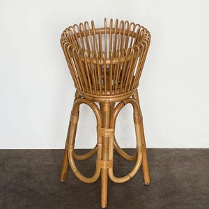 May include: A natural woven rattan plant stand with a wide, round top and a three-legged base. The stand is made of light brown rattan and has a natural finish.