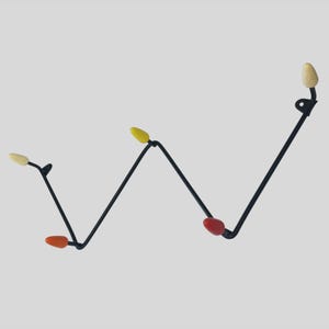 May include: A black metal wall hook with a zig-zag design. It features four coloured accents: yellow, red, and orange, with a light beige accent. The hook is designed for hanging items and adds a modern touch to any space.