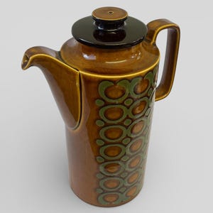 May include: A brown ceramic coffee pot with a dark brown lid and handle. The pot features a repeating pattern of green circles. The spout is curved, and the pot has a tall, cylindrical shape. A vintage kitchenware item.