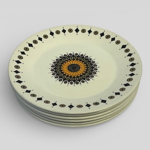 May include: A stack of six cream-coloured ceramic plates. Each plate features a central floral design in orange, brown, and black, surrounded by a border of black diamonds and gold circles. The plates are stacked neatly, showcasing their decorative pattern.