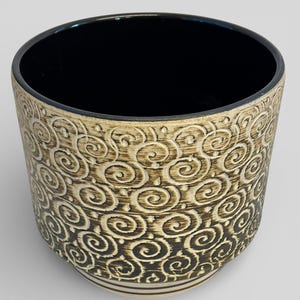 May include: A ceramic planter with a black interior and a beige exterior. The exterior features a repeating pattern of swirling, scroll-like designs in a darker brown colour. The rim is black, and the base has two horizontal lines.