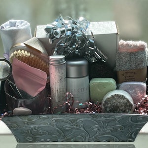 May include: A gray metal basket filled with bath and body products, including a white towel, a bath brush, a pink bag, a glass jar, two bottles, three bars of soap, a tea infuser, and a pair of gray socks. The basket is filled with pink and silver crinkle paper.