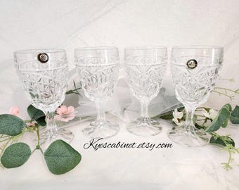 May include: Four clear crystal wine glasses with a diamond pattern. Each glass has a stem and a decorative emblem near the rim. The glasses are arranged on a white surface with floral and greenery accents.