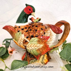 May include: A ceramic teapot shaped like a pheasant with a brown, green, and red design. The teapot has a spout and handle and is decorated with leaves and feathers.