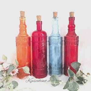 May include: Four decorative glass bottles in shades of orange, red, and blue, each with a cork stopper. The bottles have a textured design and are arranged in a row. Floral accents are in the foreground.