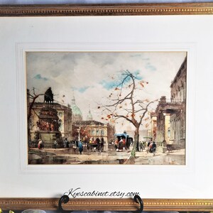 Op de afbeelding: Een ingelijst aquarel schilderij dat een straatbeeld in een Europese stad voorstelt. Het schilderij toont een standbeeld op de voorgrond, gebouwen op de achtergrond en mensen die over straat lopen. De lucht is bewolkt en de bomen zijn kaal.