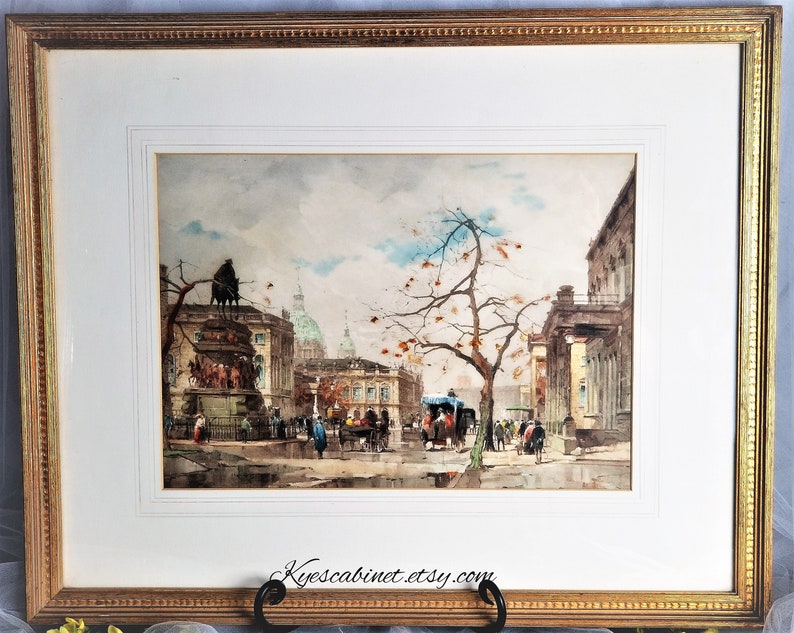 Op de afbeelding: Een ingelijst aquarel schilderij dat een straatbeeld in een Europese stad voorstelt. Het schilderij toont een standbeeld op de voorgrond, gebouwen op de achtergrond en mensen die over straat lopen. De lucht is bewolkt en de bomen zijn kaal.