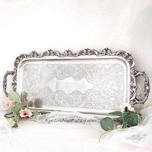 May include: A rectangular silver serving tray with ornate detailing. The tray features a decorative floral pattern and handles on each side. The border is intricately designed with a scrolling pattern. The text "Kyescabinet etsy.com" is visible.