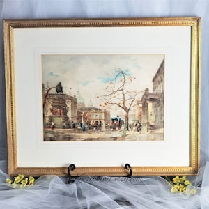 Op de afbeelding: Een ingelijst aquarel schilderij dat een stadstraat sc&egrave;ne toont met gebouwen, een standbeeld, een koets getrokken door paarden en mensen die lopen. Het schilderij is in een gouden lijst met een witte passe-partout.