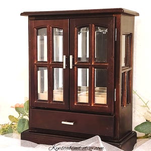 May include: A dark brown wooden jewelry cabinet with glass-paned doors and a drawer. The cabinet has a rectangular shape with a decorative top and base. The doors have silver handles, and the drawer has a single silver pull. The text "Kyescabinet etsy.com" is at the bottom.