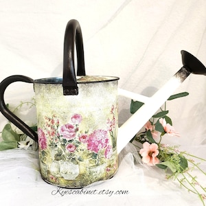 May include: Decorative watering can with a floral design featuring pink roses and hydrangeas in pots. The can has a black handle and spout, and a white spout. The website address Kyescabinet.etsy.com is printed on the bottom.