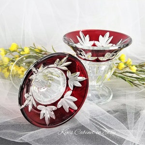 May include: Two clear glass candle holders with red bases and intricate cut glass designs. The holders have a pedestal base and a wide, shallow bowl.