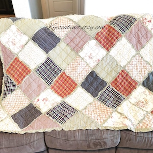 May include: A patchwork quilt with a diamond pattern in various colors, including red, white, blue, and beige. The quilt has a ruffled edge and is displayed on a brown sofa. The text "kyescabinet.etsy.com" is visible on the quilt.