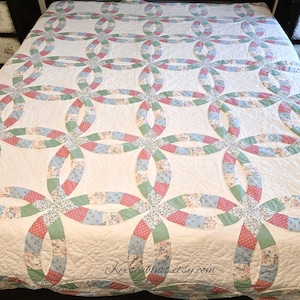 May include: A double wedding ring quilt featuring a white background with interlocking circular patterns. The rings are composed of various fabric segments in shades of sage green, light blue, and pink, featuring small floral and polka dot prints.
