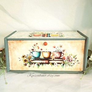 May include: A rectangular wooden bread box with a muted green frame and a cream-colored front panel. The front features a printed illustration of three teacups resting on books, surrounded by colorful wildflowers. A small orange knob is centered.