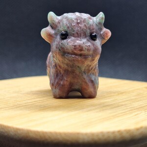 May include: A small, hand-carved stone figurine of a creature resembling a highland cow. The sculpture features a mottled texture with shades of brown, green, and grey, two small horns, and two smooth, round, black inset eyes on its face.