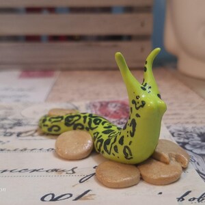 May include: A handcrafted slug figurine, featuring a vibrant yellow body with black markings. The slug is positioned on a collection of small, tan, circular pieces. The slug's body is curved, and it has two tall, curved antennae.