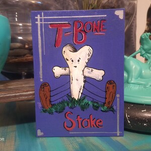 May include: A whimsical painting on a blue background features a cartoon T-bone steak with arms and a face. The words "T-Bone Stake" are written in red. The artwork is framed with silver lines and includes brown log-like posts.