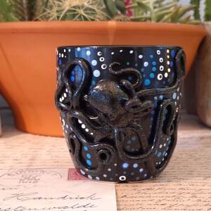 May include: Black ceramic mug with a raised octopus design. The mug features a black background with white and blue polka dots. The octopus is also black and has detailed tentacles. A unique, handmade piece.