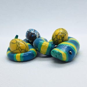 May include: A coiled snake figurine painted with blue, teal, and yellow stripes, featuring two smooth yellow stones with black crackle patterns and one blue and white speckled stone resting within its loops on a plain white background.