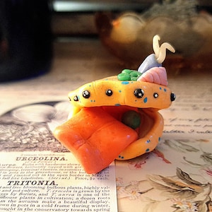 May include: A handcrafted polymer clay sculpture of a fantastical creature, with an open mouth and vibrant colours. It has orange, yellow, blue, pink, and green details, with black dot eyes. It sits on a vintage botanical print.