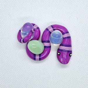 May include: A coiled purple snake figurine featuring alternating dark and light purple stripes. The snake is positioned with its body wrapped around two translucent, iridescent spherical beads and one smooth, opaque light green oval-shaped stone.