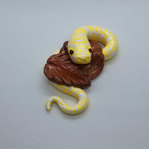 May include: A sculpted figurine of a bright yellow and white patterned snake coiled around a large, textured brown leaf. The snake has small, distinct red eyes and a smooth, glossy finish, resting in a curved position on the dark brown foliage.