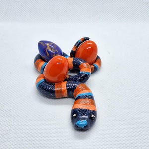 May include: A coiled snake figurine with a dark blue, glittery body featuring horizontal orange and light blue stripes. The snake has a light blue nose and small black eyes. It is positioned around two smooth, solid orange oval eggs.