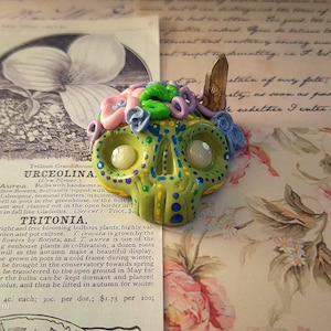 May include: A hand-painted, green skull-shaped pendant with colorful floral accents and a crystal. The skull has large, round eyes and is decorated with blue and yellow details. A unique, artistic piece.