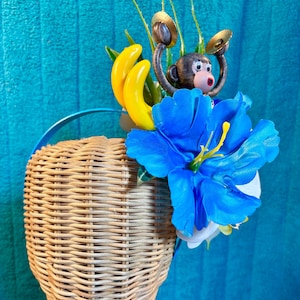 May include: A blue headband featuring a decorative arrangement of a small brown monkey figurine holding gold cymbals, two bright yellow plastic bananas, and a large vibrant blue artificial flower with green foliage accents.