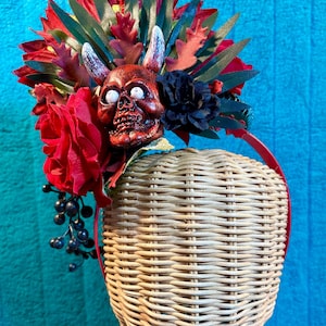 May include: A decorative headpiece featuring a red skull with white eyes, adorned with red roses, dark berries, and various leaves. The headpiece is set on a woven head form, with a red headband.