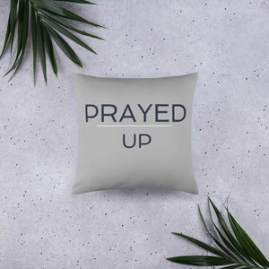 Op de afbeelding: Een grijs kussen met de tekst "PRAYED UP" in donkerblauwe letters.