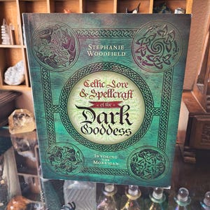 May include: A hardcover book titled Celtic Lore & Spellcraft of the Dark Goddess: Invoking the Morrigan by Stephanie Woodfield. The cover features a green textured background with black Celtic knotwork borders and four circular corner designs.