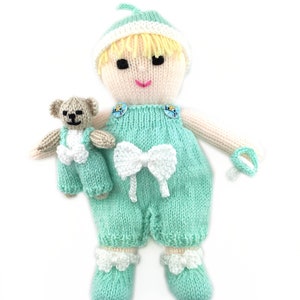 May include: A knitted doll wearing a mint green jumpsuit with a white bow and a white hat. The doll is holding a small knitted teddy bear.