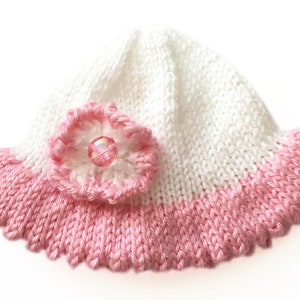 May include: A white and pink knitted baby hat with a scalloped brim. The hat features a pink flower with a clear, iridescent bead in the center. The hat is a soft, handmade item.