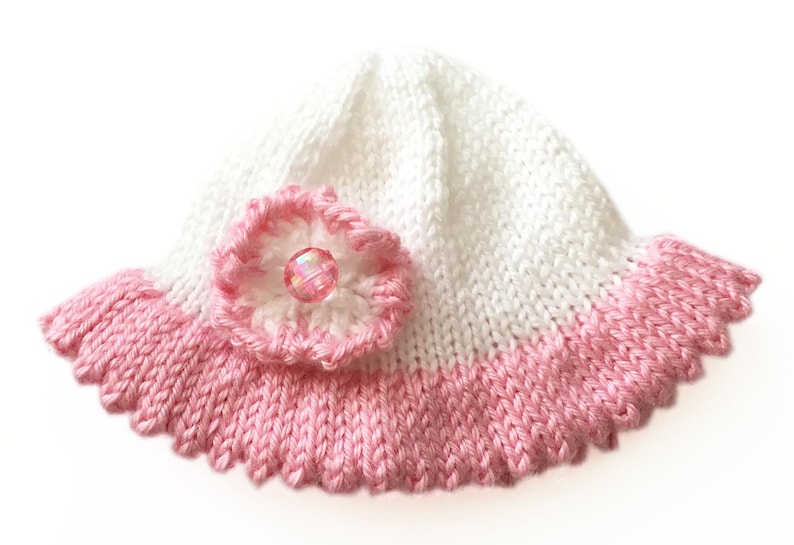 May include: A white and pink knitted baby hat with a scalloped brim. The hat features a pink flower with a clear, iridescent bead in the center. The hat is a soft, handmade item.